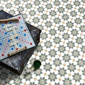 May include: A Scrabble board with letter tiles spelling out words like "AGAIN" and "PRawn" sits on a dark, textured box. A small green glass is next to the box. The background features a patterned tile floor with floral designs in white, grey, and tan.