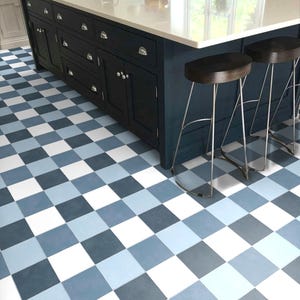 Könnte beinhalten: Eine Kücheninsel mit weißer Arbeitsplatte und dunkelblauen Schränken. Zwei braune Holzstühle mit silbernen Beinen stehen vor der Insel. Der Boden hat ein Schachbrettmuster in Blau- und Weißtönen.