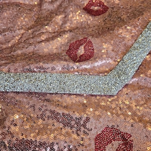 Puede incluir: Una tela de lentejuelas en oro rosa con un patrón repetitivo de motivos de labios en lentejuelas rojo oscuro. El material está acentuado con un borde ancho en forma de V de lentejuelas plateadas densas y brillantes.