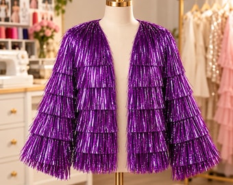 May include: A vibrant purple jacket featuring multiple horizontal tiers of metallic fringe. The garment has long sleeves, an open front design, and a collarless neckline, displayed on a mannequin to showcase the layered texture.