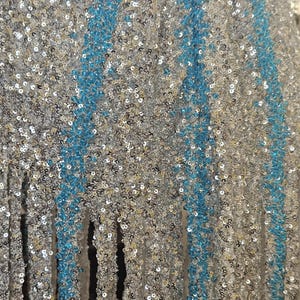 May include: A fabric surface densely covered in shimmering silver sequins, featuring vertical stripes of bright turquoise sequins. The material has a textured, reflective appearance with a fringe-like bottom edge where the sequined fabric hangs down.
