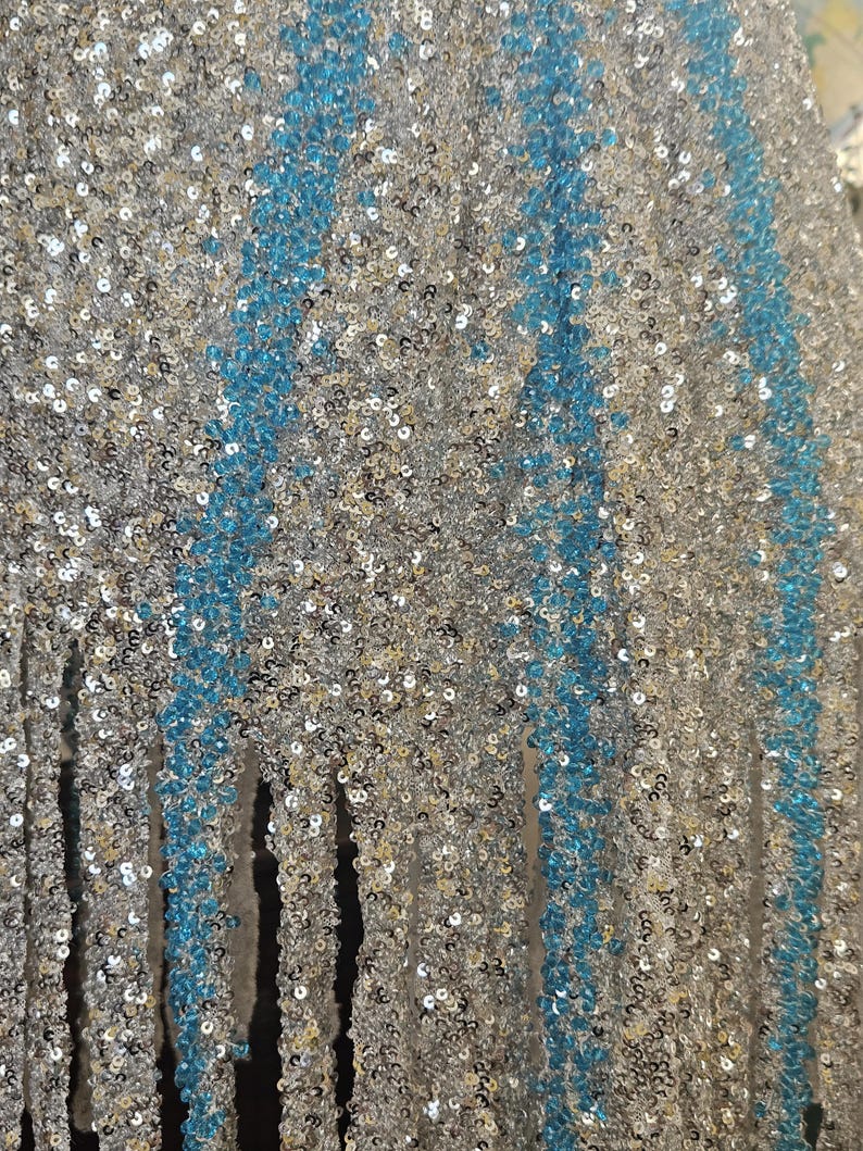 May include: A fabric surface densely covered in shimmering silver sequins, featuring vertical stripes of bright turquoise sequins. The material has a textured, reflective appearance with a fringe-like bottom edge where the sequined fabric hangs down.