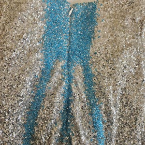 May include: A sleeveless dress covered in shimmering silver sequins with a prominent vertical stripe of bright blue sequins running down the center. The garment features a V-neckline, thin shoulder straps, and a concealed zipper closure at the back.