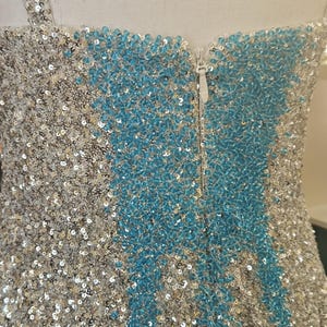 May include: A sleeveless garment covered in shimmering silver and bright blue sequins. The design features a central vertical panel of blue sequins flanked by silver sequins, with a white zipper closure running down the center of the back.