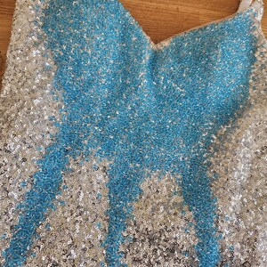 May include: A sleeveless top covered in shimmering sequins featuring a V-neckline. The garment displays a central pattern of bright blue sequins contrasted against a background of silver sequins, with white fabric straps visible at the shoulders.