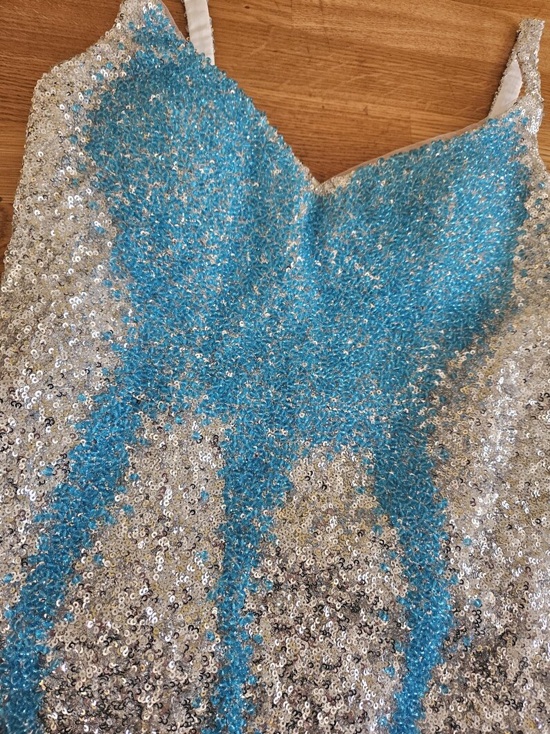 May include: A sleeveless top covered in shimmering sequins featuring a V-neckline. The garment displays a central pattern of bright blue sequins contrasted against a background of silver sequins, with white fabric straps visible at the shoulders.