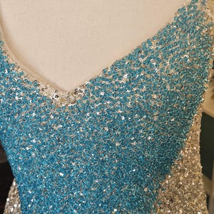May include: A sleeveless dress featuring a V-neckline, fully covered in shimmering sequins. The garment displays a distinct color-blocked pattern with a central panel of bright turquoise sequins flanked by silver sequins on the sides.