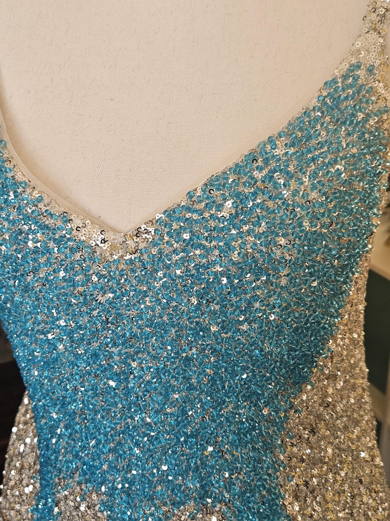 May include: A sleeveless dress featuring a V-neckline, fully covered in shimmering sequins. The garment displays a distinct color-blocked pattern with a central panel of bright turquoise sequins flanked by silver sequins on the sides.