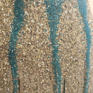 May include: A garment featuring a dense pattern of shimmering silver sequins interspersed with vertical stripes of vibrant turquoise beads. The lower edge of the piece is finished with long, dangling fringe strands made of matching silver sequins.