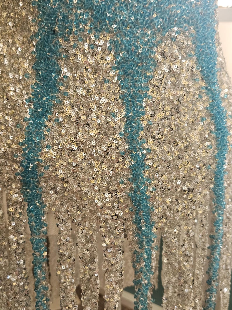 May include: A garment featuring a dense pattern of shimmering silver sequins interspersed with vertical stripes of vibrant turquoise beads. The lower edge of the piece is finished with long, dangling fringe strands made of matching silver sequins.