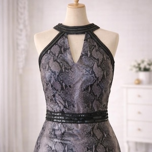 Puede incluir: Vestido sin mangas con estampado de piel de serpiente en tonos grises y negros. El vestido presenta un cuello halter con un corte de ojo de cerradura y una banda negra adornada en la cintura. El vestido se exhibe en un maniquí.