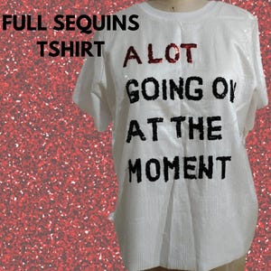 May include: A white short-sleeved t-shirt covered in small white sequins. The front features the text A LOT GOING ON AT THE MOMENT in black sequins, with the word A in red sequins. The shirt is displayed on a mannequin against a red glitter background.