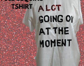 Puede incluir: Camiseta blanca con diseño de lentejuelas. La camiseta tiene el texto "A LOT GOING ON AT THE MOMENT" en letras negras.