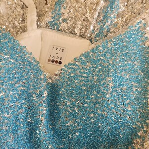 May include: A garment covered in shimmering silver and bright turquoise sequins with a white interior lining. A white fabric label inside the neckline features the text IVIE + JADE with four circular icons underneath the brand name.