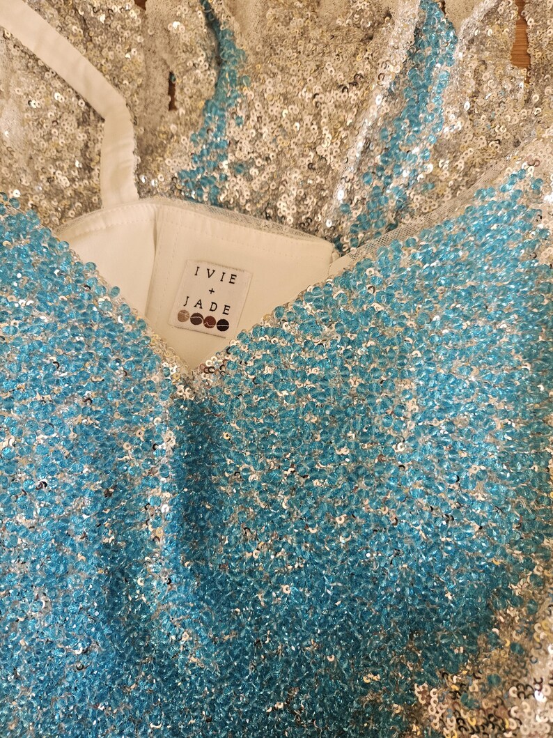 May include: A garment covered in shimmering silver and bright turquoise sequins with a white interior lining. A white fabric label inside the neckline features the text IVIE + JADE with four circular icons underneath the brand name.