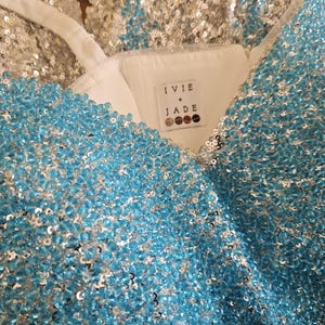 May include: A sleeveless garment featuring a white interior lining and a label reading IVIE + JADE. The exterior is densely covered in shimmering light blue beads and silver sequins, creating a textured, reflective surface with thin white straps.