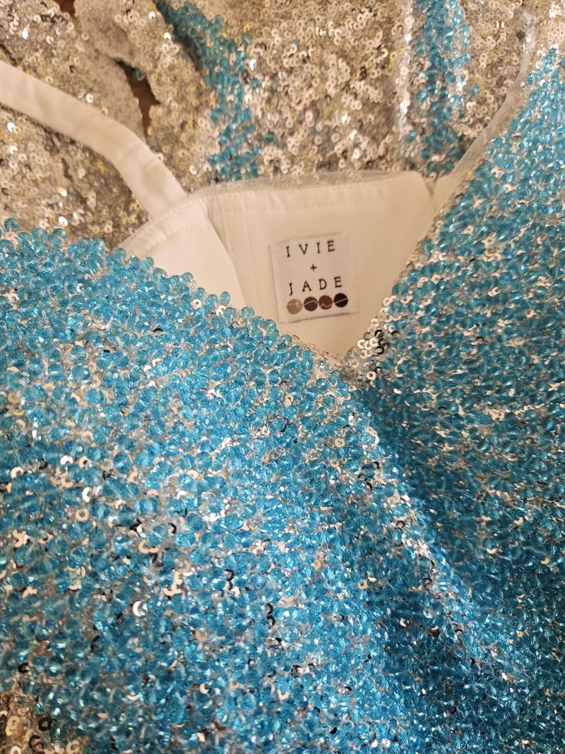 May include: A sleeveless garment featuring a white interior lining and a label reading IVIE + JADE. The exterior is densely covered in shimmering light blue beads and silver sequins, creating a textured, reflective surface with thin white straps.