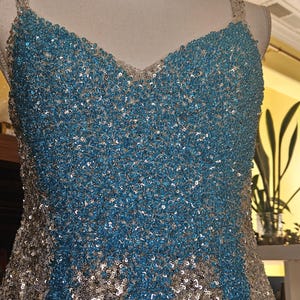 May include: A sleeveless dress featuring a V-neckline and thick straps, fully covered in a dense pattern of shimmering turquoise blue and silver sequins. The bodice displays a central panel of turquoise sequins flanked by silver side panels.