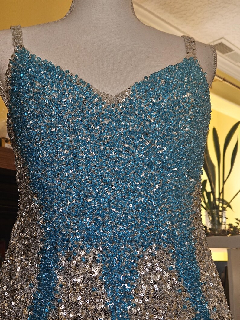 May include: A sleeveless dress featuring a V-neckline and thick straps, fully covered in a dense pattern of shimmering turquoise blue and silver sequins. The bodice displays a central panel of turquoise sequins flanked by silver side panels.