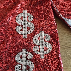 Puede incluir: Una tela roja vibrante cubierta de lentejuelas densas y brillantes. Tres símbolos de dólar distintos, hechos de pequeños diamantes de imitación claros y reflectantes, están dispuestos en un patrón triangular sobre la superficie roja texturizada.