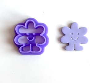 May include: Two purple flower-shaped cookie cutters. One is a cutter with a smiling face, arms, and legs. The other is a flower-shaped cookie with a smiling face. Both are on a white background.