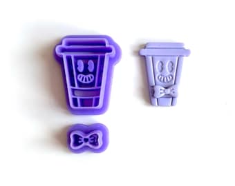 May include: Three purple cookie cutters in the shape of a cartoon coffee cup with a smiling face and a bow tie. The set includes two cup shapes and a bow tie shape. The cookie cutters are made of plastic.