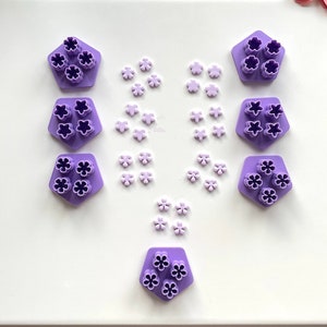 Puede incluir: Cortadores de galletas en forma de flor de color morado dispuestos en un patrón simétrico sobre una superficie blanca. También hay pequeñas decoraciones en forma de flor de color morado esparcidas alrededor de los cortadores.