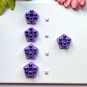 Może przedstawiać: Cztery fioletowe plastikowe foremki do ciasteczek w kształcie motyla, każda z sześcioma wycięciami w kształcie motyla, są ułożone pionowo na białym tle. Cztery małe białe foremki do ciasteczek w kształcie motyla są ułożone pionowo po prawej stronie fioletowych foremek.