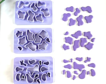Op de afbeelding: Drie sets paarse plastic koekjesvormen in de vorm van abstracte organische vormen. Elke set is in een rij op een wit oppervlak gerangschikt. Naast elke set bevinden zich de uitgesneden vormen van paarse fondant.
