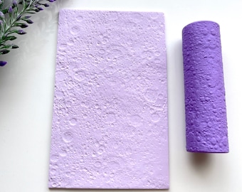 May include: A light purple textured rectangular mold and a cylindrical roller. The mold and roller have a pattern of small craters, resembling a lunar surface. The items are on a white surface, with a sprig of purple flowers in the upper left corner.