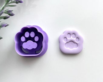 May include: A purple paw print cookie cutter and a paw print-shaped cookie. The cookie cutter is a darker shade of purple and has a detailed paw print design. The cookie is a lighter shade of purple.