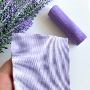 May include: A sheet of light purple textured faux leather with a woven pattern. The sheet is held in a hand.