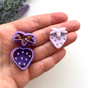 Op de afbeelding: Een set van twee paarse plastic koekjesvormen in de vorm van aardbeien. De vormen hebben een gedetailleerd ontwerp met kleine hartjes voor de zaadjes.
