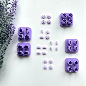 Può includere: Un set di timbri in argilla polimerica viola con varie icone meteorologiche, tra cui nuvole, pioggia, sole, fulmini e vento. I timbri sono disposti in file su una superficie bianca.