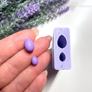 Puede incluir: Dos pequeños objetos con forma de huevo, moteados y de color lavanda, se sostienen cerca de un molde lavanda con dos cavidades en forma de huevo. Los objetos están sobre una superficie blanca, con un fondo borroso de flores moradas.