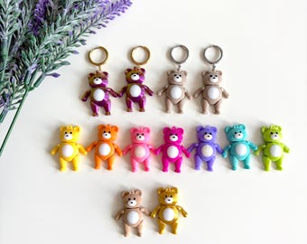 May include: A set of 12 colorful teddy bear keychains. The keychains are made of plastic and have a shiny finish. The bears are in various colors, including pink, purple, blue, green, yellow, and orange. The keychains are arranged in two rows, with the top row featuring four bears and the bottom row featuring eight bears.