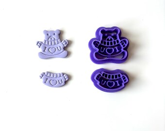 May include: Four purple cookie cutters in the shape of a teddy bear and a banner. The bear and banner have the words "I U" and a heart. The cookie cutters are made of plastic.