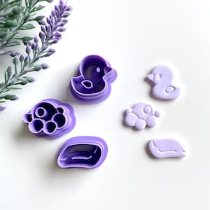 Puede incluir: Un juego de cinco cortadores de galletas de plástico morado con forma de pato, nube, bañera, burbujas y jabón.