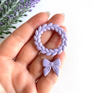 May include: A set of two lavender-colored polymer clay embellishments. One is a wreath with a circle shape and the other is a bow.