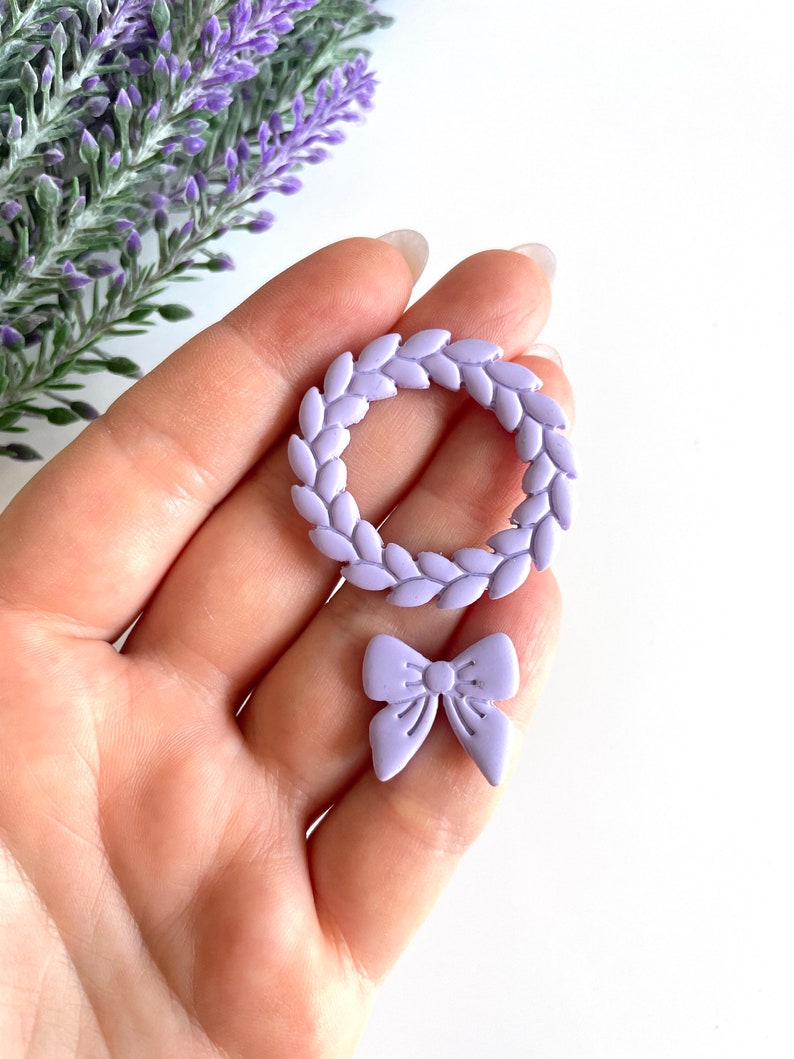 May include: A set of two lavender-colored polymer clay embellishments. One is a wreath with a circle shape and the other is a bow.