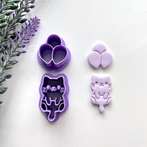 May include: Purple plastic cookie cutters in the shape of a cat and a bunch of balloons. The cat cutter has a black outline and the balloon cutter has a white outline.
