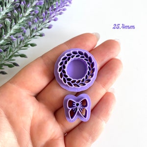 May include: Two purple plastic cookie cutters, one shaped like a wreath and the other shaped like a bow. The wreath cutter is 2.54 inches in diameter.