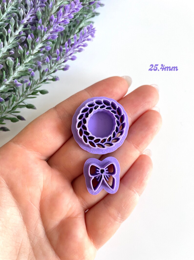 May include: Two purple plastic cookie cutters, one shaped like a wreath and the other shaped like a bow. The wreath cutter is 2.54 inches in diameter.