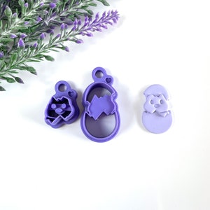May include: Three purple plastic cookie cutters in Easter shapes. The cutters depict a decorated egg, a chick hatching from an egg, and a small egg with a heart-shaped top. They are on a white surface with lavender sprigs, ideal for baking.