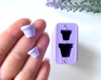May include: A light purple silicone mold with two flower pot impressions, and two small, light purple flower pot miniatures. The flower pots have a ribbed design. The background includes lavender-colored floral sprigs.