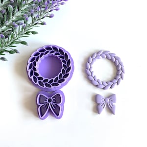 May include: A set of three purple plastic cookie cutters. The cutters are shaped like a wreath, a wreath with leaves, and a bow.