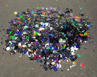 May include: A pile of iridescent glitter with a mix of black, silver, and colorful hexagonal shapes. The glitter reflects light, creating a shimmering effect. The glitter is scattered on a dark gray surface.