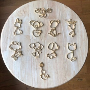 May include: A set of ten wooden animal and object cutouts, including a turtle, koala, frog, dog, cat, and a ball. The cutouts are all gold in color and are arranged on a light brown wooden surface.