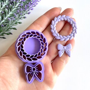 May include: Three purple plastic cookie cutters: a wreath, a bow, and a smaller wreath. The cutters are on a hand.