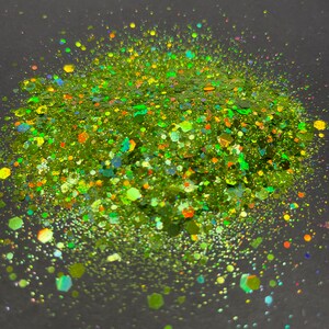 May include: Green, yellow and orange glitter with iridescent sparkle. The glitter is scattered on a dark grey background.